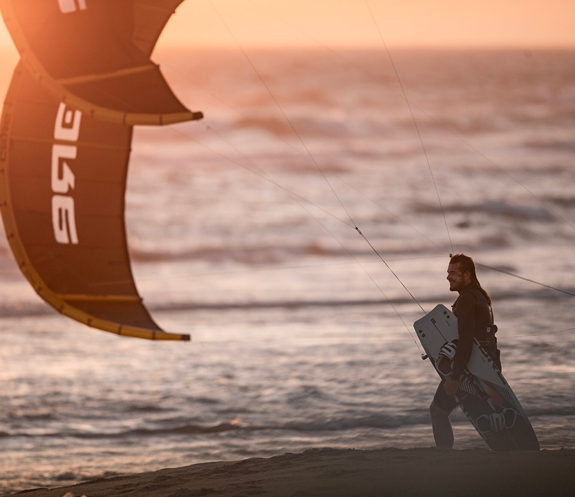 Praticante caminhando na praia ao pôr do sol, carregando kite e prancha Core
